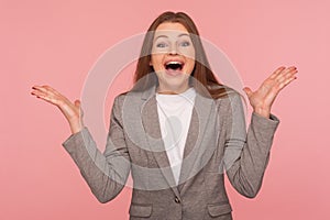 Portrait of surprised young brunette woman in business suit keeping hands raised and shouting in amazement