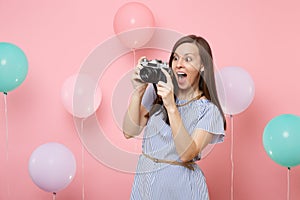 Portrait of surprised young happy woman in blue dress take pictures on retro vintage photo camera on pink background