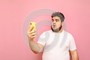 Portrait of surprised young overweight man on pink background looks into smartphone screen with shocked face wearing white t-shirt