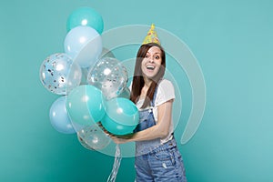 Portrait of surprised young woman in denim clothes, birthday hat celebrating, holding colorful air balloons isolated on