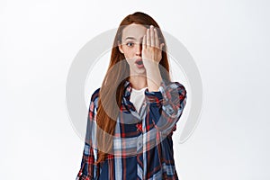 Portrait of surprised young woman gasp, say wow, cover half of face, look with one eye at camera in awe, standing in