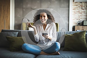 Portrait of a suspicious woman holding phone looking at you sitting on a sofa in the living room of a house