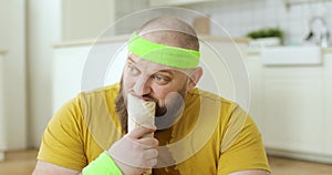 Portrait of sweaty fat man in yellow wet sportswear is eating danar in kitchen.
