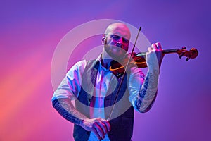 Portrait of talented young man, solo performer playing violin against gradient purple background in neon light