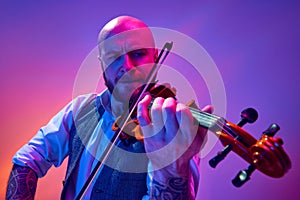 Portrait of talented young man, solo performer playing violin against gradient purple background in neon light