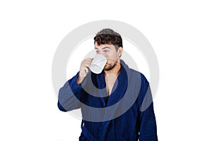Portrait of tardy young man wears blue bathrobe holding cup of coffe unable to wake up in time to get to work, isolated on white