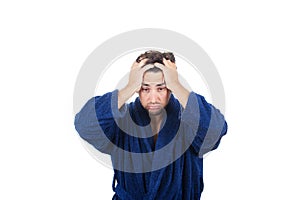 Portrait of tardy young man wears blue bathrobe holding hands to head, unable to wake up in time to get to work, isolated on white