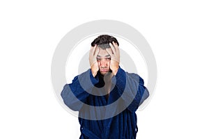 Portrait of tardy young man wears blue bathrobe holding hands to head, unable to wake up in time to get to work, isolated on white