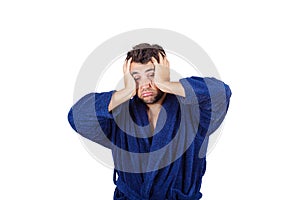 Portrait of tardy young man wears blue bathrobe holding hands to head, unable to wake up in time to get to work, isolated on white