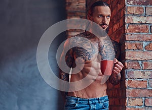 Portrait of a tattoed shirtless male with a stylish haircut and beard, drinks morning coffee, leaning against a brick