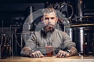 Portrait of a tattooed hipster male with stylish beard and hair in shirt sitting at the bar counter with glass of beer