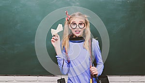 Portrait of a teen female student. School teenager girl holding backpack and paper plane, standing against blackboard.