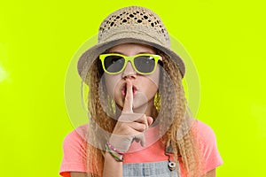 Portrait of teen girl in sunglasses and straw hat with a forefinger near the lips, showing silence sign, secret gesture, yellow