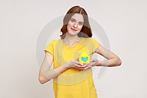 Portrait of teen girl in yellow t-shirt holding in hands near heart little paper house looking at camera with pleasant smile,