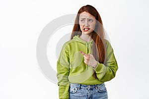 Portrait of teen redhead girl cringes, frowns and looks reluctant at something nasty or disgusting, ponting finger left