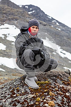 Portrait of Caucasian teenage boy dressing warm hiking outfit for mountaineering, young man sitting on big stone in mountains