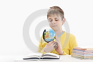 Portrait of teenage boy sitting at desk and look closely at the globe. Schoolboy does homework at the table. Isolated on white