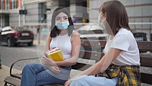 Portrait of teenage Caucasian girl with black hair in coronavirus face mask sitting on bench on city street talking with