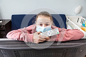 Portrait of teenage Caucasian girl with tv remote control in hands, wearing medical mask, tired to stay at home