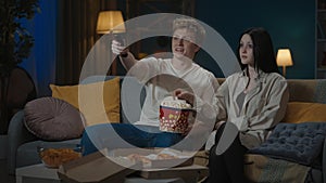 Portrait of teenage couple spending leisure time. Boy and girl sitting on the sofa eating popcorn and watching movie.