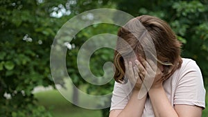 Portrait of a teenage girl standing on the street in a park on a Summer day, covering her face with her hands, crying. A sad child