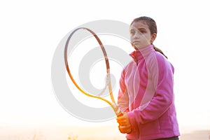 Portrait of teenage girl is training tennis skill outside court, in field at sunset. Wearing pink sweater and holding racket.