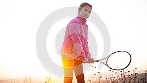 Portrait of teenage girl is training tennis skill outside court, in field at sunset. Wearing pink sweater.