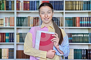 Portrait of teenage student with books in hands looking at camera in school library