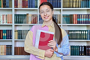 Portrait of teenage student with books in hands looking at camera in school library