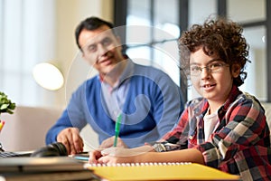 Portrait of teenaged latin boy in glasses smiling at camera, sitting at the desk together with his father and doing