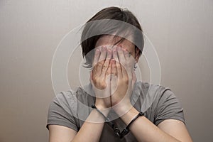 Portrait of a teenager in handcuffs covering his face with his hands on a gray background, medium plan. Juvenile delinquent, crimi