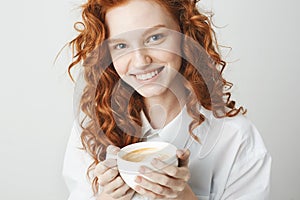 Portrait of tender redhead girl with freckles smiling holding cup looking at camera. White background.