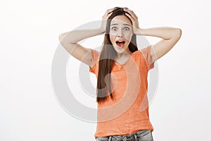 Portrait of terrified shocked beautiful european woman in orange t-shirt, holding hands on head and yelling from horror