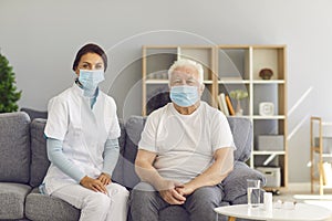 Portrait of therapeutist and aged white-haired man, both wearing medical face masks, sitting on couch at home.