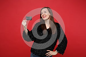 Portrait of thinkful young woman in black fur sweater looking up, holding credit bank card isolated on bright red