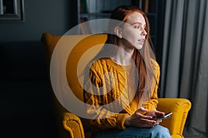 Portrait of thinking redhead young woman using mobile phone sitting in yellow armchair, pensive looking away.