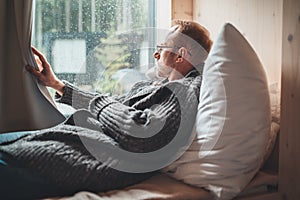 Portrait of thinking sad Middle-aged man in eyeglasses dressed open cardigan lying on cozy bed next to window looking out street