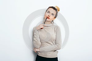 Portrait of Thinking woman with appealing smile, having hair bun in sweater isolated on white background holding pen and have a pe