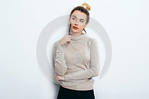 Portrait of Thinking woman with appealing smile, having hair bun in sweater isolated on white background holding pen and have a pe