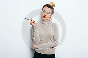 Portrait of Thinking woman with appealing smile, having hair bun in sweater isolated on white background holding pen and have a pe