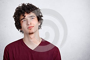 Portrait of thinking young man with dark curly hair, stands with thoughtful facial expression, dresses maroon shirt. Student