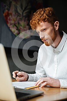 Portrait of thinking young readhead man looking at laptop at workplace