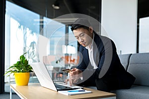 Portrait thoughtful Asian businessman working on a laptop computer at a modern office desk. Confident Focused pensive man