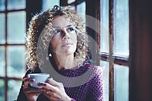 Portrait of thoughtful attractive adult woman at home holding a cup of tea and looking outside the windows - Pretty curly young