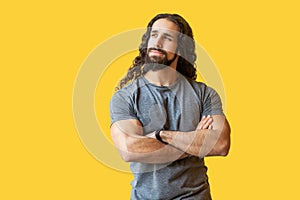 Portrait of thoughtful bearded young man with long curly hair in grey tshirt standing, crossed hands, looking away and thinking