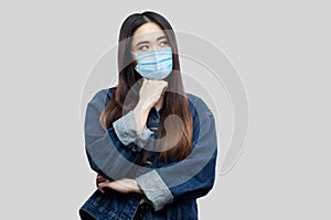 Portrait of thoughtful beautiful asian young woman with surgical medical mask in blue denim jacket standing, confused looking away