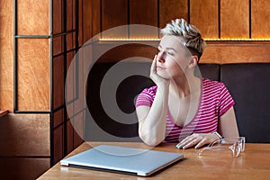 Portrait of thoughtful beautiful young bussineswoman with blonde short hair in pink t-shirt is sitting in cafe, finishing work and