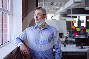 Portrait of thoughtful caucasian businessman in lowered face mask looking out of window