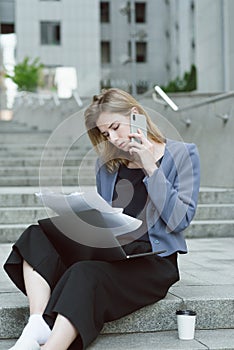 Portrait of thoughtful caucasian female office worker checking documents talking via phone. Busy employer sitting on stairs near