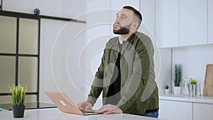 Portrait of thoughtful concentrated Caucasian man standing in kitchen with laptop. Bearded young inspired guy thinking